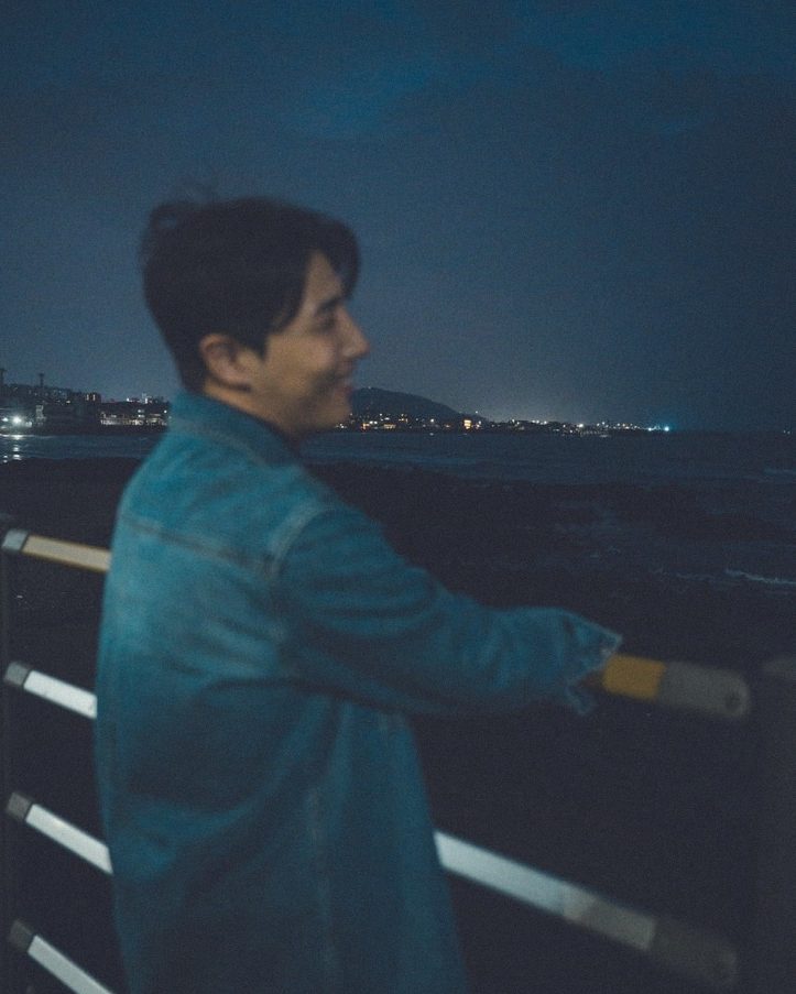 Leaning on a railing by the sea at night, city lights behind